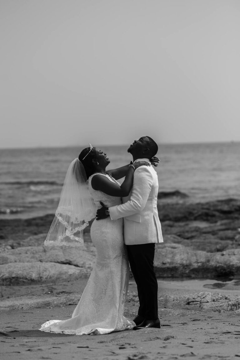 Side view of black and white happy young African American bride and groom embracing and smiling while standing on sandy beach near sea on wedding day